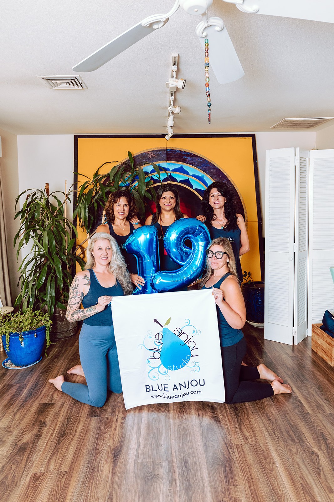 Five Blue Anjou teachers holding blue 19 balloons beside the studio banner