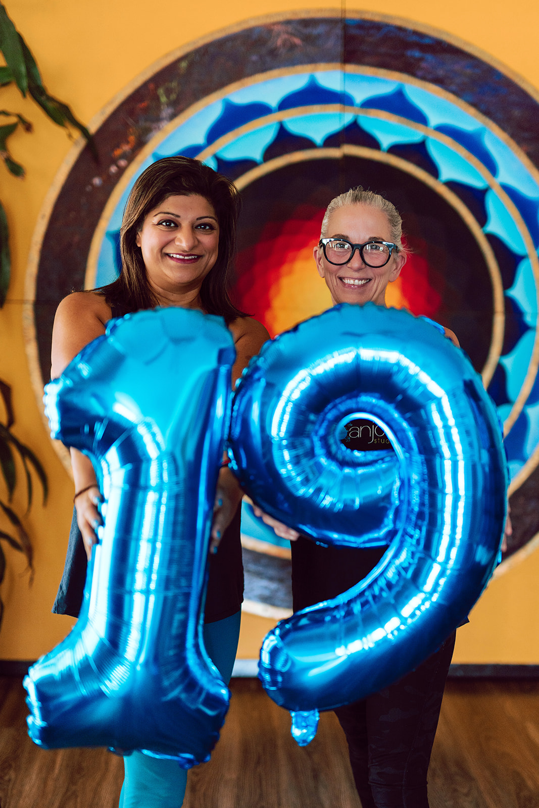 Two Blue Anjou teachers holding 19 balloons in front of the mandala mural