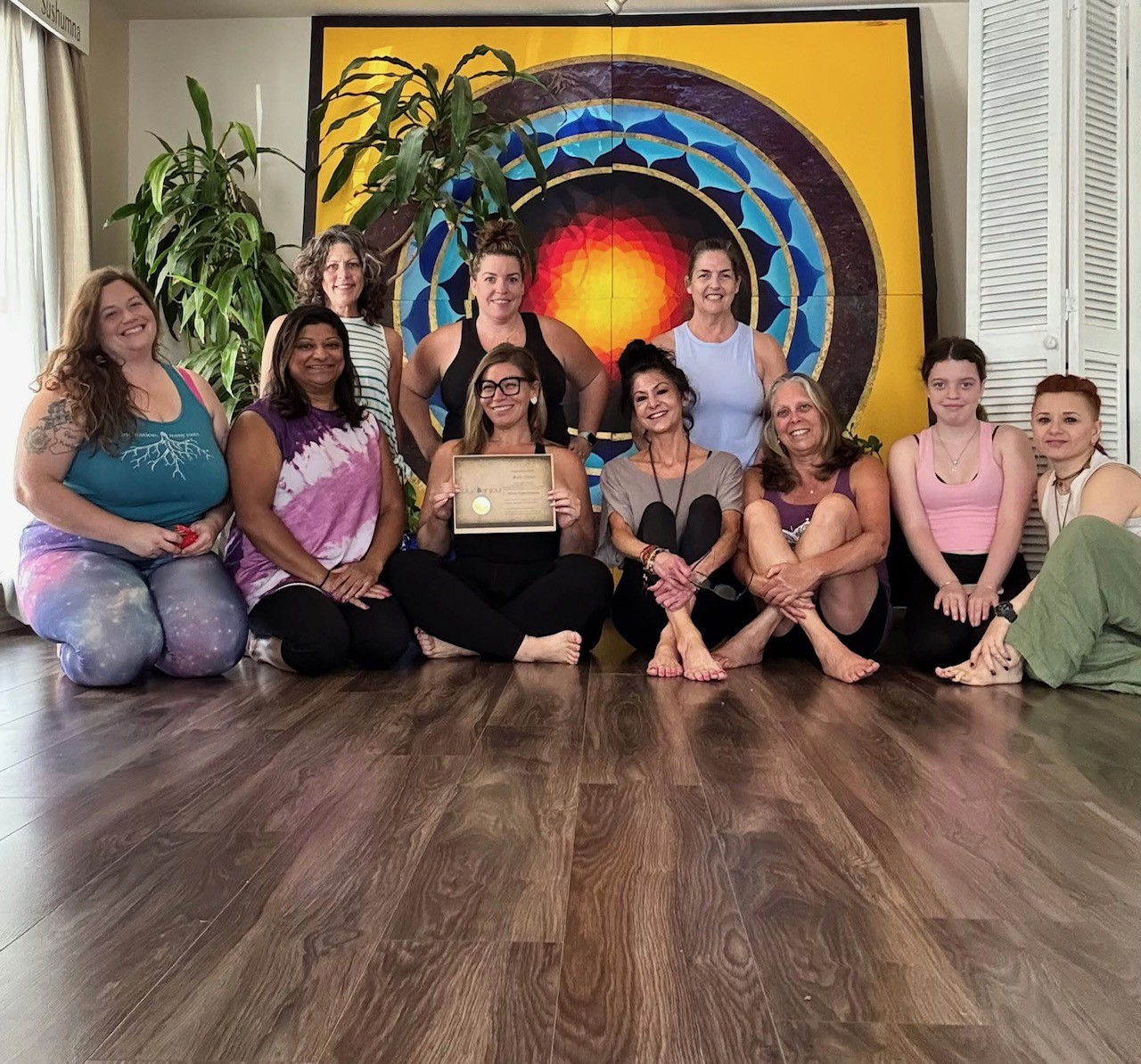 Blue Anjou teacher training group gathered in front of the studio mandala wall